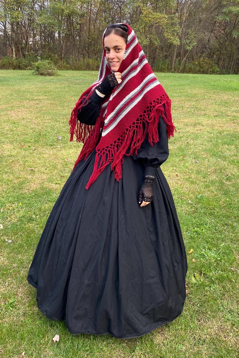 Puede incluir: Una persona vestida con un vestido negro y un chal rojo, blanco y gris con flecos. El chal est&aacute; drapeado sobre la cabeza y los hombros. El vestido tiene mangas largas y una falda amplia. Se usan guantes de encaje negros.