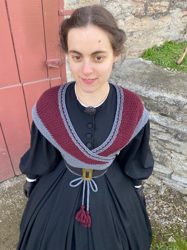 May include: A woman in a black dress with a burgundy and grey knitted shawl. The shawl is draped over the shoulders and around the waist, secured with a belt. The dress has long sleeves and a high neckline.
