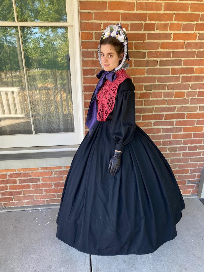 May include: A woman wearing a black dress with a large skirt, a red crocheted shawl, and a white bonnet with purple flowers.