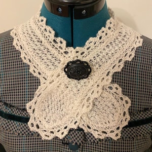 May include: Cream-colored, crocheted collar with a scalloped edge and a black oval brooch. The collar is draped over a teal dress form, showcasing the intricate openwork design and vintage aesthetic. The brooch features a floral design.