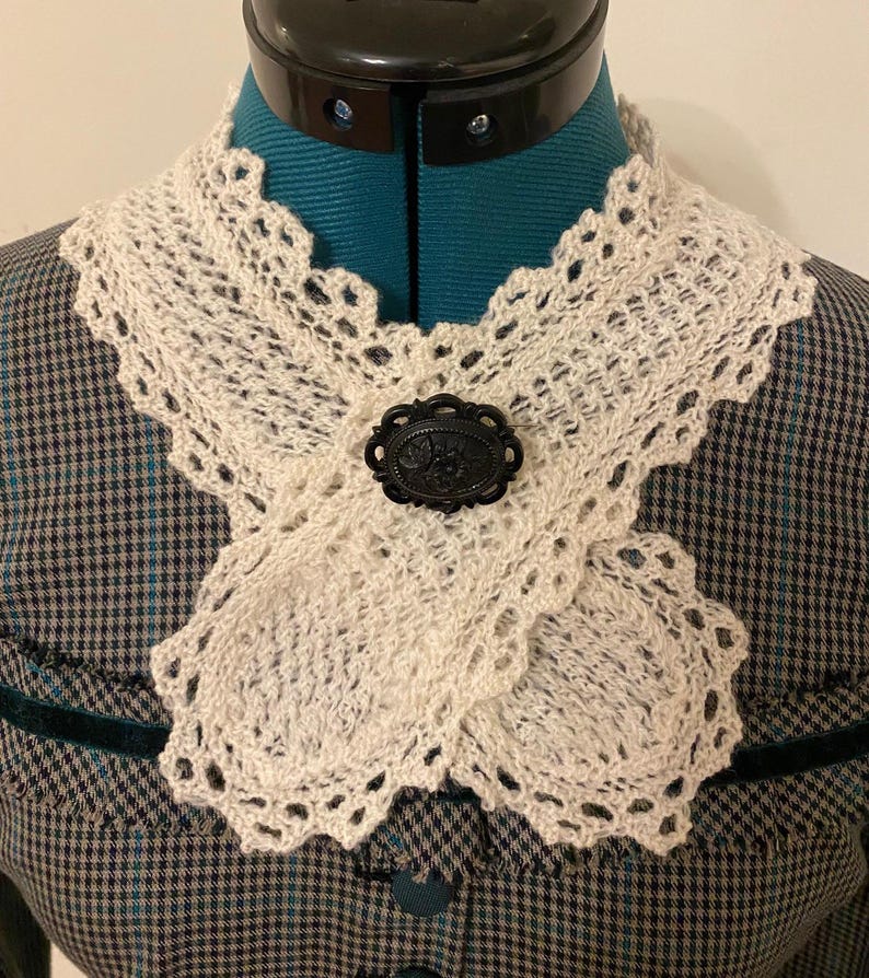 May include: Cream-colored, crocheted collar with a scalloped edge and a black oval brooch. The collar is draped over a teal dress form, showcasing the intricate openwork design and vintage aesthetic. The brooch features a floral design.