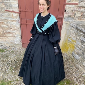May include: A black Victorian dress with a full skirt and long sleeves. The dress has a turquoise and black shawl collar and a matching belt. The sleeves have white cuffs, and the person is wearing black gloves. The background includes a red door and a stone wall.