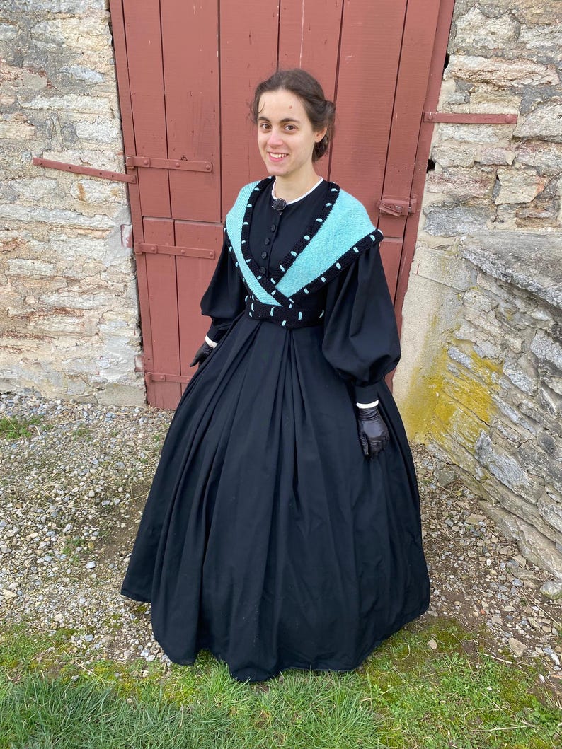 May include: A black Victorian dress with a full skirt and long sleeves. The dress has a turquoise and black shawl collar and a matching belt. The sleeves have white cuffs, and the person is wearing black gloves. The background includes a red door and a stone wall.