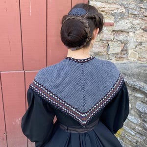 May include: A woman in a black dress with a grey knitted shawl. The shawl features a decorative border with white, brown, and purple accents. Her hair is styled in a bun with a decorative hair comb.