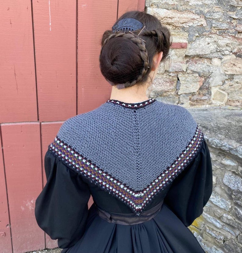 May include: A woman in a black dress with a grey knitted shawl. The shawl features a decorative border with white, brown, and purple accents. Her hair is styled in a bun with a decorative hair comb.