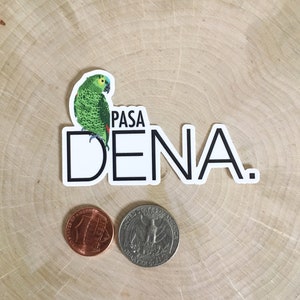 May include: A sticker featuring a green parrot with blue and yellow accents perched above the text "PASA DENA." A penny and a quarter are next to the sticker on a wooden surface.