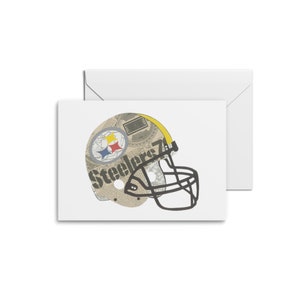 May include: A white card with a black and white illustration of a football helmet with the word "Steelers" printed on it. The helmet is made to look like it is made of newspaper.