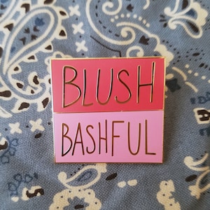 May include: A gold-coloured pin with the words "BLUSH" in pink and "BASHFUL" in light pink.