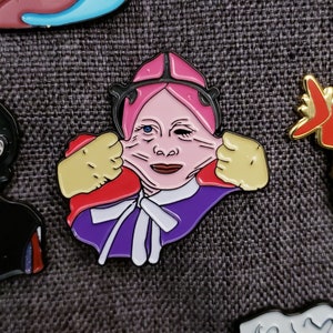 May include: A pink and purple enamel pin featuring a cartoon character with a surprised expression. The character has pink hair, a purple dress, and is holding their cheeks with their hands.