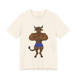 May include: Off-white t-shirt with a brown, muscular bull graphic. The bull is wearing blue shorts and has its arms crossed. The design is in a geometric style. The shirt has a crew neck and short sleeves.