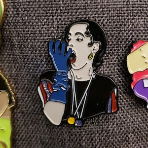 May include: Enamel pin depicting a person with black hair, wearing a black top and blue glove, with their mouth open. The pin has gold, white, red, and blue accents. A collectible item.