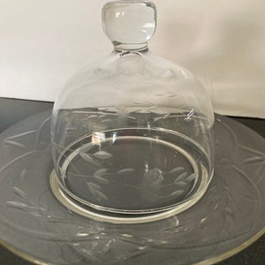 May include: A clear glass cloche with a knob on top sits on a matching clear glass plate with an etched design. The cloche and plate are both made of glass and have a delicate, elegant look.