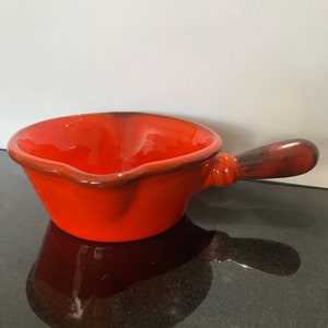 May include: A red ceramic sauce pot with a brown handle. The pot has a rounded shape and a pouring spout.