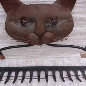 May include: A rusty metal cat sculpture with a rake attached to the bottom. The cat's eyes are made of metal and have a spiral design. The rake has 12 tines.