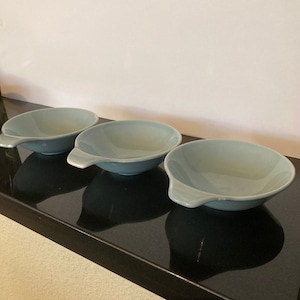 May include: Three light blue ceramic bowls with a handle on the side. The bowls are on a black surface.