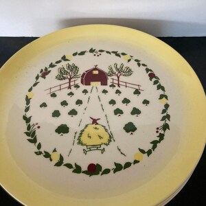 May include: A yellow rimmed white ceramic plate with a farmhouse scene. The scene features a red barn, green trees, a hay wagon, and a wreath of green leaves with red berries.