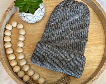 Puede incluir: Un gorro de punto gris con motas de hilo blanco. El gorro está sobre una bandeja de madera con una pequeña suculenta en maceta y una guirnalda de cuentas de madera.