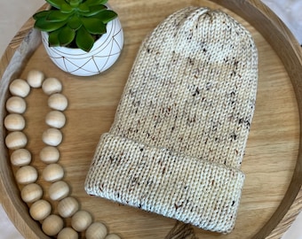 Puede incluir: Un gorro de punto color crema con motas marrones, doblado en el borde. El sombrero se exhibe en una bandeja de madera con una pequeña suculenta en una maceta geométrica blanca y dorada, y una hebra de cuentas de madera.