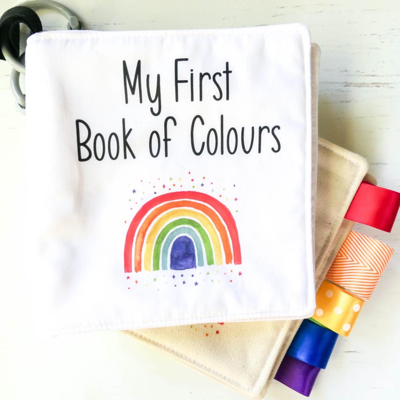 May include: A white cloth book with the title "My First Book of Colours" and a rainbow illustration. The book has colorful ribbon tags attached to the side.