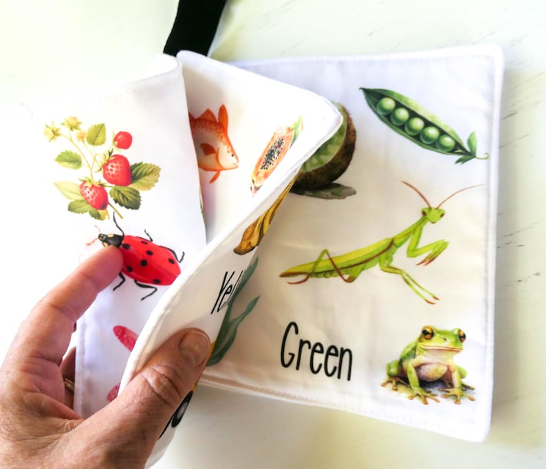 May include: A white fabric book with colorful illustrations of a ladybug, strawberries, a fish, a papaya, an avocado, a praying mantis, and a frog. The book has the words "Yellow" and "Green" printed on it.