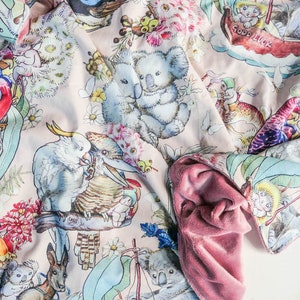 May include: A pink fabric baby blanket with a whimsical design featuring koalas, parrots, kookaburras, and other Australian animals. The blanket has a pink plush backing.