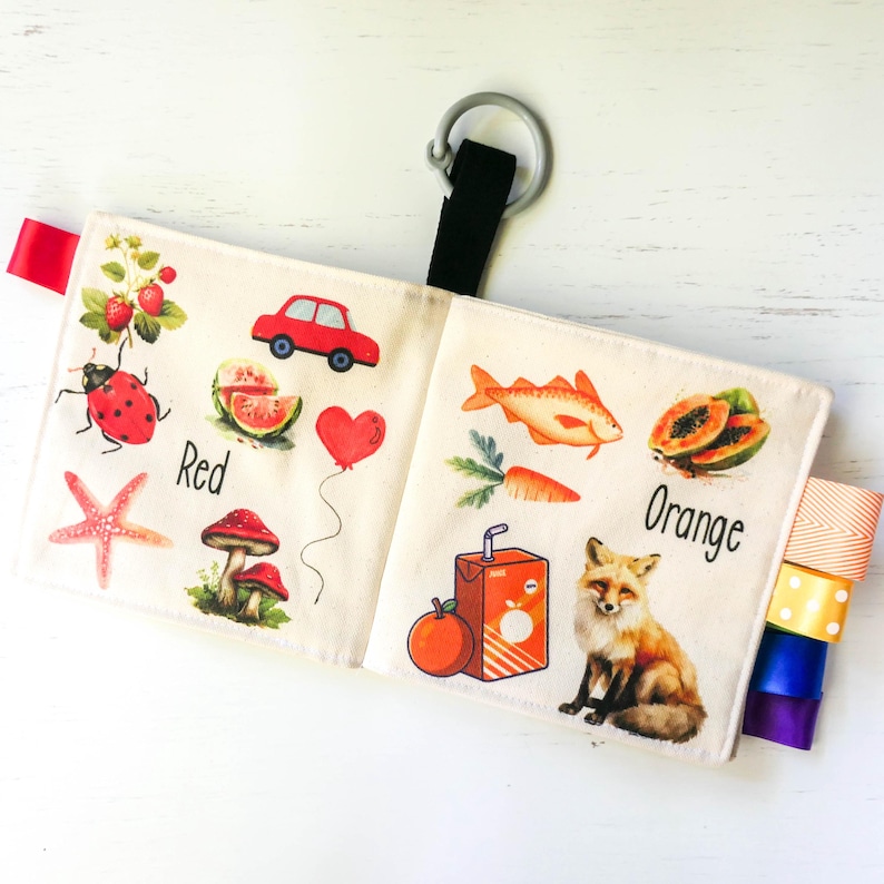 May include: A soft cloth book for babies and toddlers with colorful illustrations of objects and animals. The book features the colors red and orange with images of a ladybug, a car, a watermelon, a heart, a starfish, a mushroom, a fish, a carrot, a papaya, an orange, and a fox. The book has colorful ribbons attached to the side.