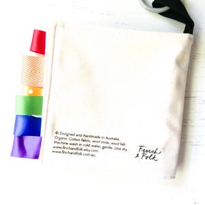 May include: A white canvas bag with a black strap and rainbow ribbon trim. The bag has the text "Designed and Handmade in Australia. Organic Cotton fabric, wool inner, wool felt. Machine wash in cold water, gentle. Line dry. www.finchandfolk.etsy.com www.finchandfolk.com.au" and "Finch & Folk" printed on it.