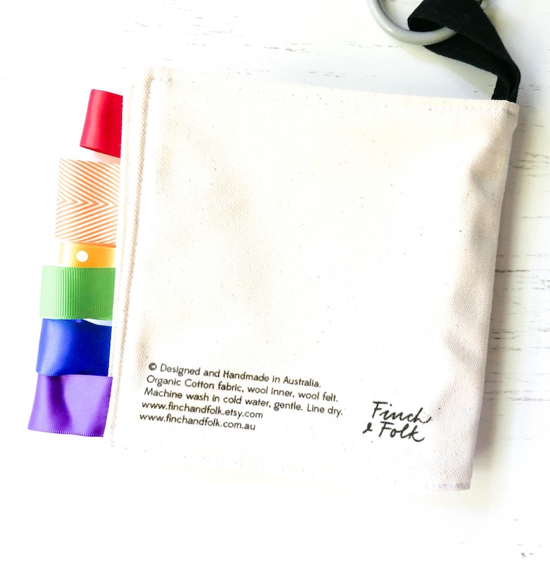 May include: A white canvas bag with a black strap and rainbow ribbon trim. The bag has the text "Designed and Handmade in Australia. Organic Cotton fabric, wool inner, wool felt. Machine wash in cold water, gentle. Line dry. www.finchandfolk.etsy.com www.finchandfolk.com.au" and "Finch & Folk" printed on it.