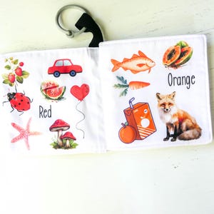 May include: A soft fabric book for babies and toddlers with colorful illustrations of objects and the words "Red" and "Orange". The book features a ladybug, a car, a watermelon, a mushroom, a fish, a carrot, a fox, and an orange juice box.