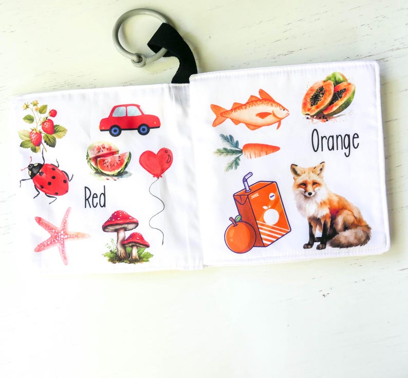 May include: A soft fabric book for babies and toddlers with colorful illustrations of objects and the words "Red" and "Orange". The book features a ladybug, a car, a watermelon, a mushroom, a fish, a carrot, a fox, and an orange juice box.