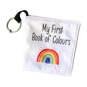 May include: A white fabric book with a rainbow and the text "My First Book of Colours" printed on the front. The book has a black and grey plastic ring attached to the top.