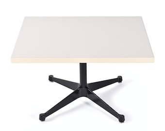 May include: A square table featuring a smooth, off-white top with light-colored edges. The table is supported by a central black metal column that connects to a four-pronged base, with each prong ending in a small, circular, metallic foot.