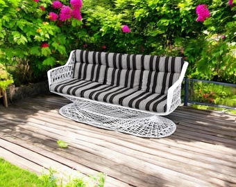 May include: A white outdoor swing bench with a striped black and white cushion. The bench is made of a woven material and sits on a wooden deck. Lush green foliage and pink flowers are in the background.