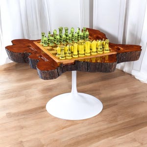 May include: A unique chess table with a live edge wood design. The table features a brown, organic-shaped top with a built-in chessboard. The chess pieces are yellow and green. The table is supported by a white, tulip-style base.