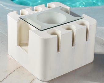 May include: A white, cube-shaped outdoor table with a glass top and recessed areas for holding glasses. The table has a modern design and is likely made of a durable, weather-resistant material.