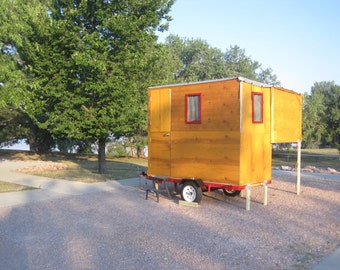 May include: A small, wooden, yellow-painted cabin on wheels with two windows and a door. The cabin is sitting on a gravel driveway with a grassy area in the background.