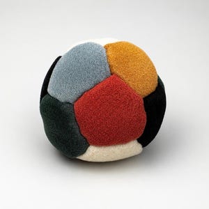 May include: A colorful, plush ball with a geometric design. The ball features various colors, including red, orange, blue, green, black, and white. The soft, textured surface suggests a toy or decorative item.