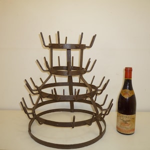 以下が含まれることがあります： 複数の突起が付いた、3段のヴィンテージメタルボトル乾燥ラック。赤いキャップとラベルが付いたワインボトルがラックの隣にあります。ラックは濃い茶色の金属製です。