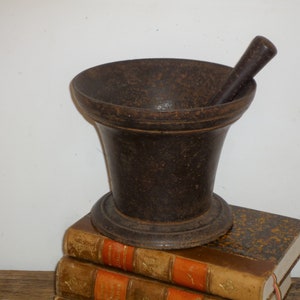 Peut inclure: Un mortier et un pilon bruns foncés. Le mortier a un bol évasé et une base arrondie. Le pilon est cylindrique. L'ensemble est posé sur deux livres anciens aux couvertures usées.