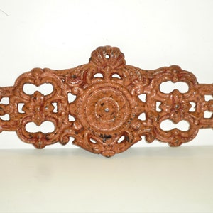 May include: A decorative cast iron wall plaque with an intricate design in a reddish-brown color. The plaque features a central circular motif surrounded by floral and scrollwork.