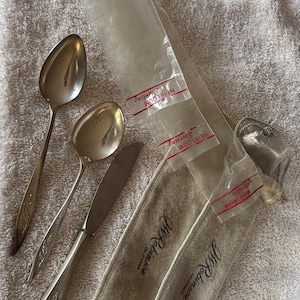 May include: Two silver spoons and one silver knife with decorative handles, accompanied by two beige fabric storage pouches labeled J.W. Robinson California and three clear plastic sleeves labeled Heirloom Sterling.
