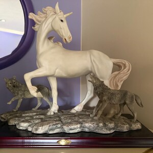 May include: A white unicorn figurine with a spiraled horn, standing with two gray wolves. The wolves are positioned at the unicorn's feet, one howling. The base is a dark wood with a stone-like texture.