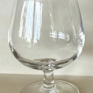 May include: A clear, stemmed brandy snifter glass. The glass has a rounded bowl, a short stem, and a wide, circular base. The glass is transparent, allowing light to pass through, and is designed for holding and swirling brandy or other spirits.