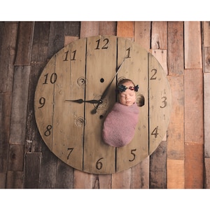 May include: A newborn baby wrapped in a mauve blanket, wearing a floral headband, positioned on a large, rustic wooden clock face. The clock hands point to approximately 9:10. The background is a wooden plank wall.