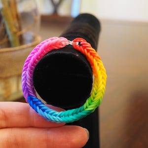 May include: A circular bracelet constructed from small, interconnected rubber bands in a vibrant rainbow gradient pattern. The colors transition from pink and red to orange, yellow, green, blue, and purple, secured by a clear plastic S-clip.