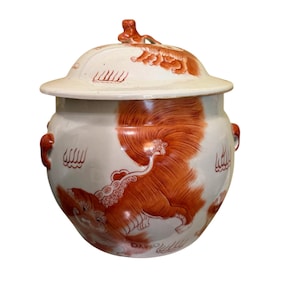 Puede incluir: Un jarrón de cerámica blanco con tapa, con ilustraciones naranjas de una criatura mítica. El jarrón tiene forma redonda con dos asas y una tapa con un adorno decorativo. El diseño es de estilo tradicional.