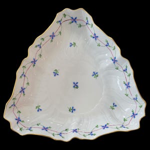 Puede incluir: Plato de porcelana blanca con borde festoneado y ribete dorado. El plato está decorado con detalles florales azules y verdes y una vid rosa. El plato tiene forma triangular.