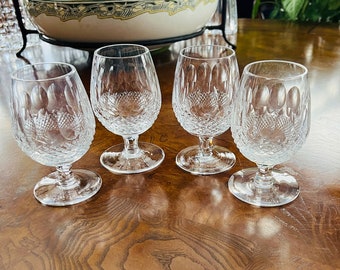 Puede incluir: Cuatro copas de coñac de cristal transparente con un diseño tallado. Las copas tienen forma de cuenco redondeado, un tallo corto y una base circular. Están dispuestas sobre una superficie de madera pulida.