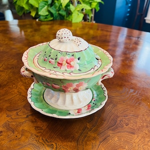 Könnte beinhalten: Eine grüne und weiße Porzellan-Zuckerdose mit Deckel und passendem Unterteller. Die Schale ist mit rosa und roten Blumen und grünen Blättern verziert. Der Deckel hat einen kleinen Knopf in Form einer Erdbeere.