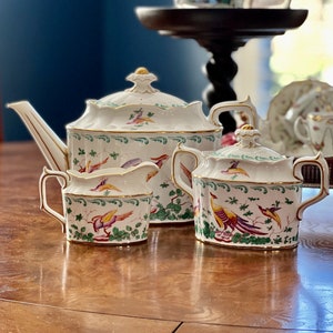May include: A white porcelain tea set with a green, gold and red floral pattern featuring colourful birds. The set includes a teapot, creamer and sugar bowl.
